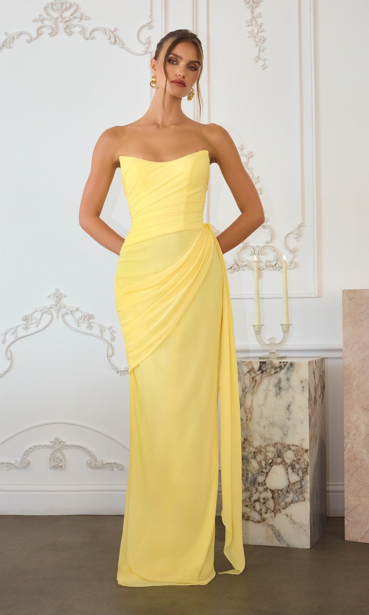 A woman in the Amoris T726 long prom dress, light yellow and strapless with draped chiffon details, stands in an elegant room featuring ornate white walls and marble accents.